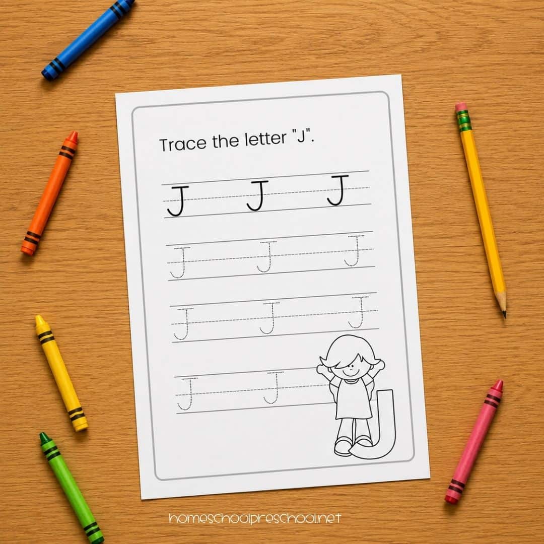 Capital letter tracing worksheet for the letter J showing solid letter models and dotted tracing rows on a wood table surrounded by colorful crayons and a yellow pencil.