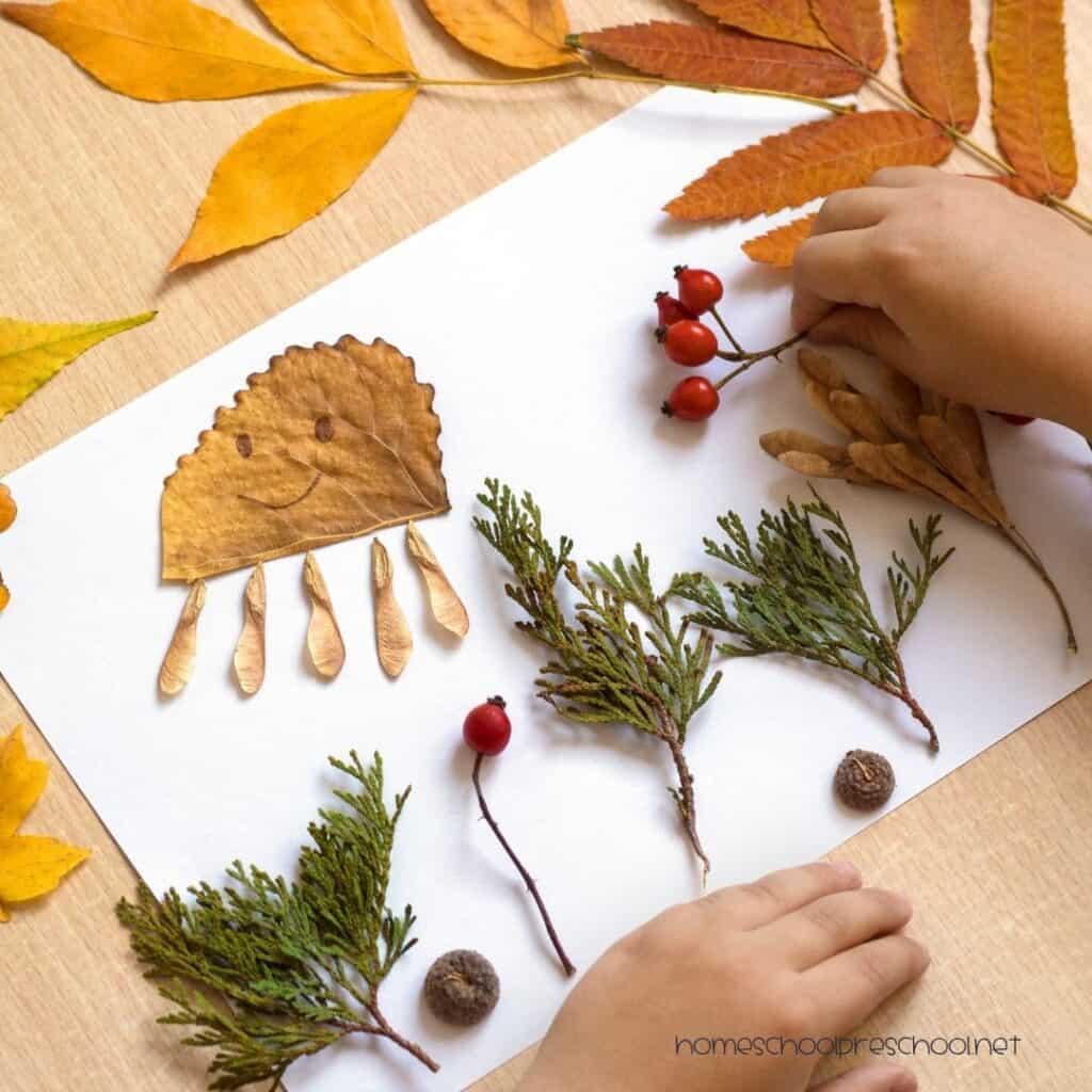 Preschooler creating fall nature art with leaves, berries, seed pods, acorns, and evergreen sprigs on white paper.
