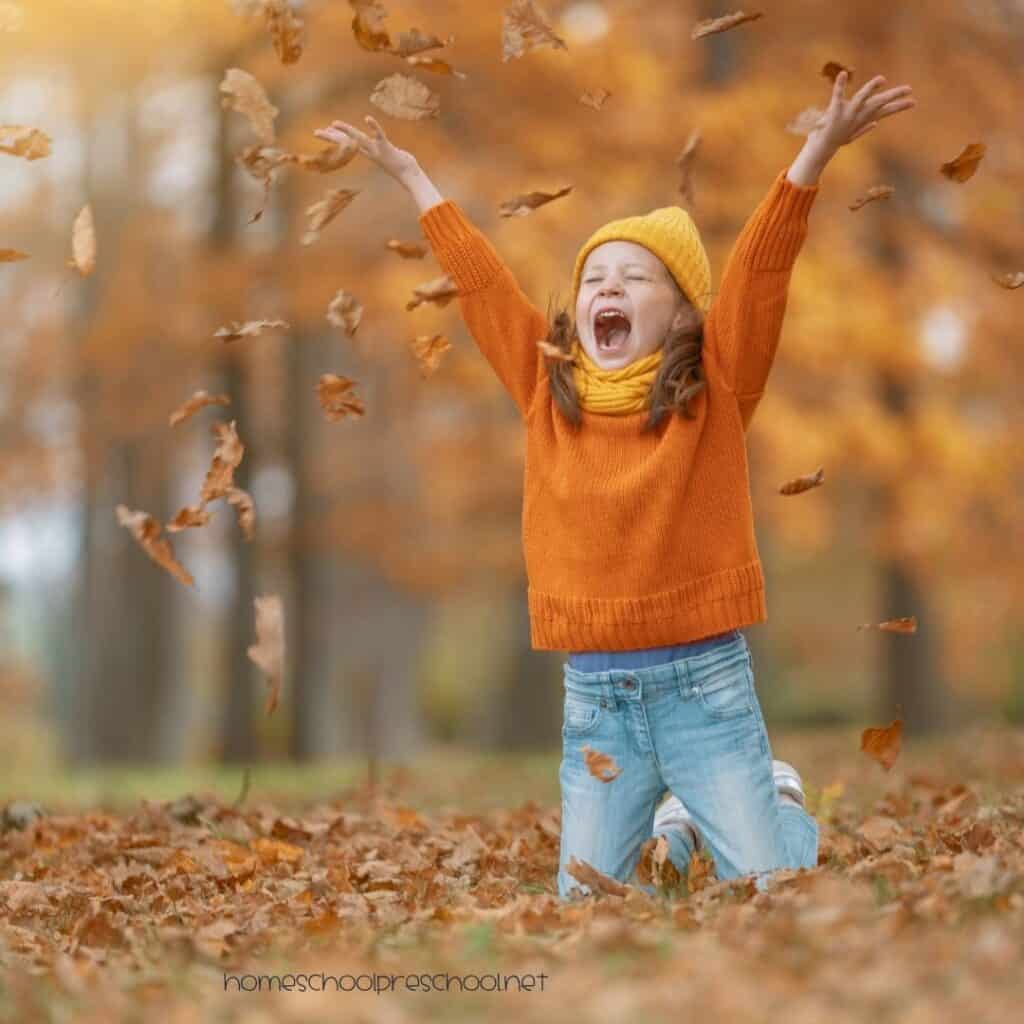 Preschooler wearing an orange sweater and yellow hat tossing fall leaves in the air outdoors.