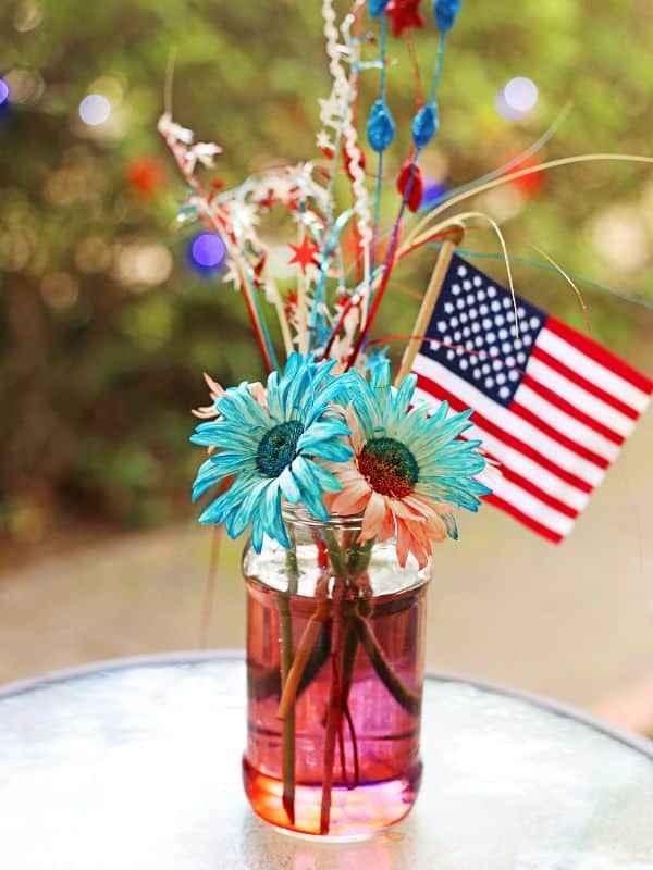 Patriotic color changing flowers with red and blue tinted petals displayed in a jar with an American flag and festive decorations.
