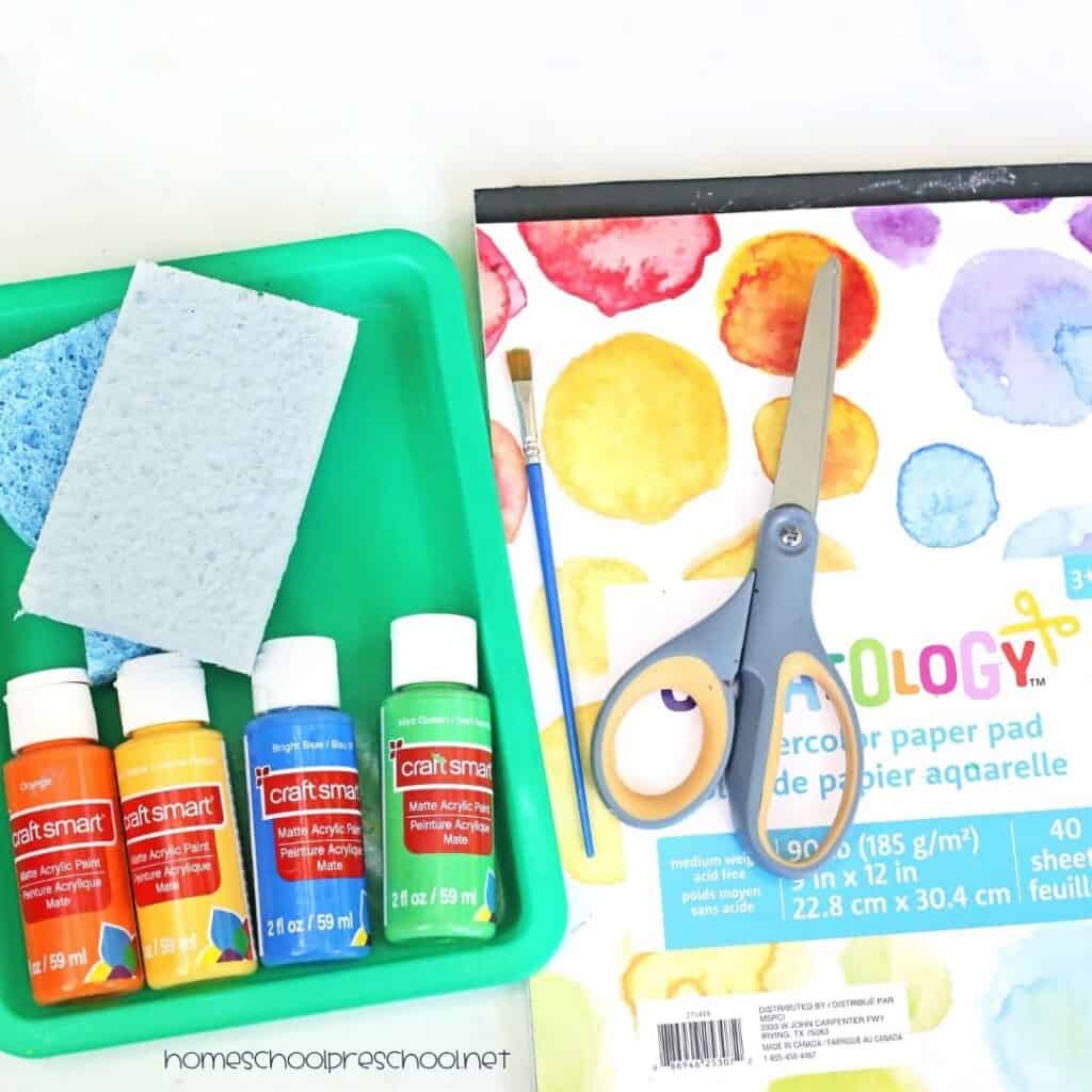 Supplies for a sponge-painted popsicle craft including sponges, colorful paint, a paintbrush, scissors, and watercolor paper arranged on a tabletop.