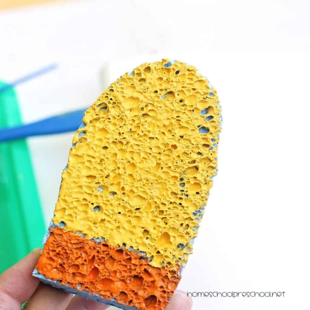 Yellow and orange paint applied to a popsicle-shaped sponge before stamping it onto paper for a summer craft activity.