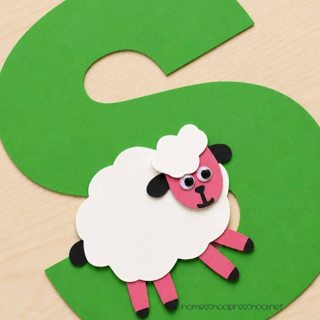 Close-up of a finished letter S sheep craft featuring a white paper sheep with a pink face attached to a large green letter S for preschool alphabet learning.