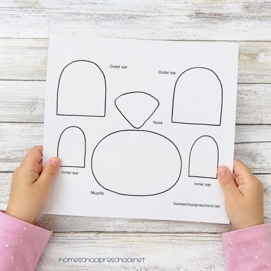 Child holding a printable bear craft template with labeled pattern pieces for the ears, muzzle, and nose used to make a bear paper plate craft.