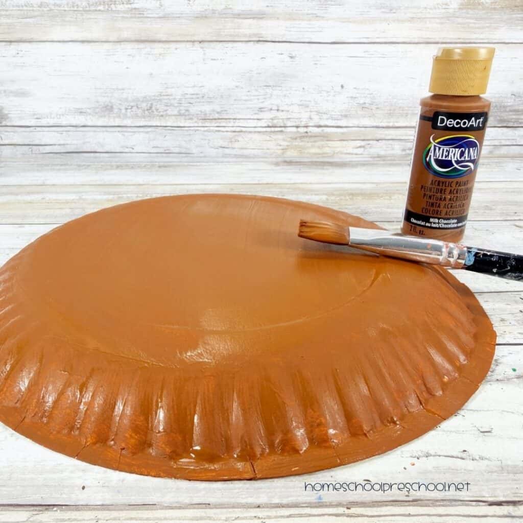 Brown paper plate painted with acrylic paint for a bear paper plate craft, with a paintbrush and brown craft paint beside it.