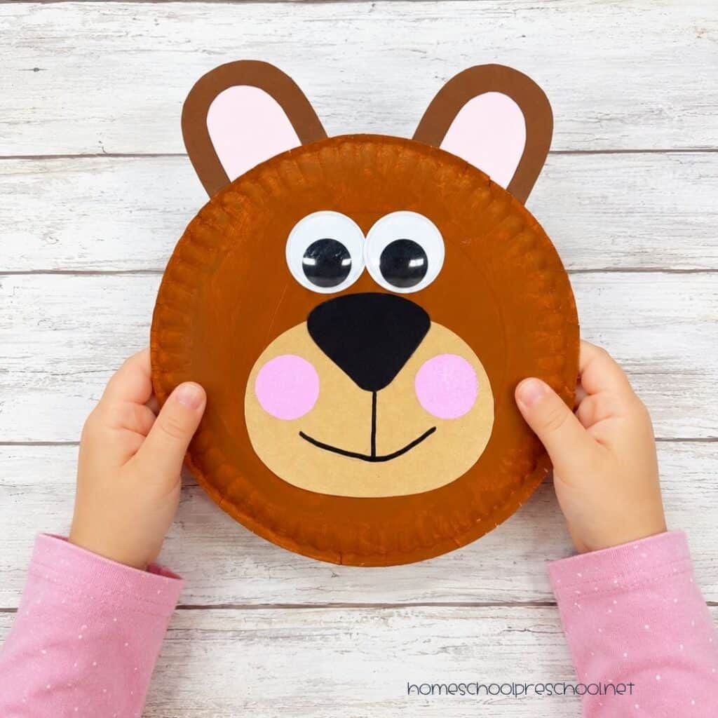 Child holding a finished bear paper plate craft with googly eyes, pink cheeks, and a smiling face made from a painted paper plate.