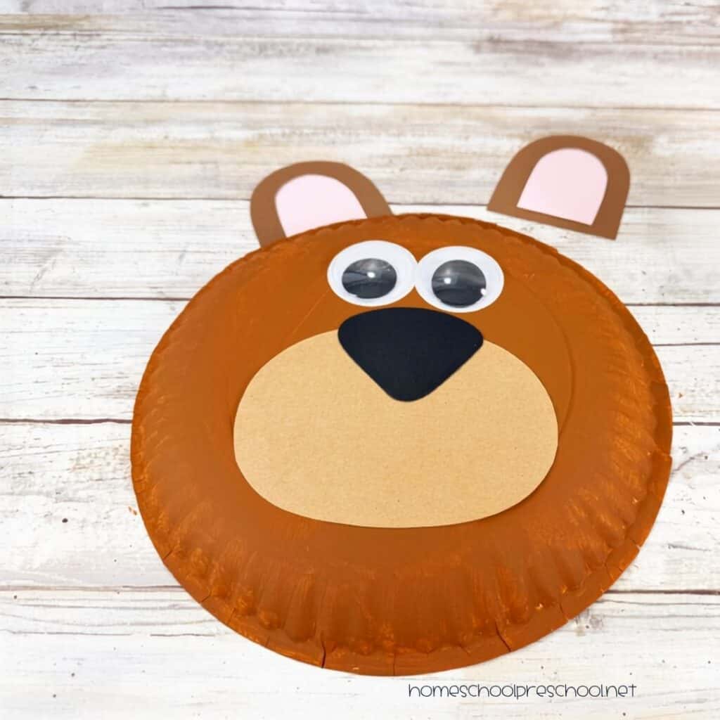 Bear paper plate craft with the ears, muzzle, nose, and googly eyes glued onto a painted paper plate before adding the final details.