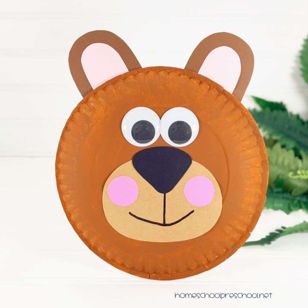 Finished bear paper plate craft made with a painted brown paper plate, cardstock ears and muzzle, googly eyes, pink cheeks, and a smiling face.