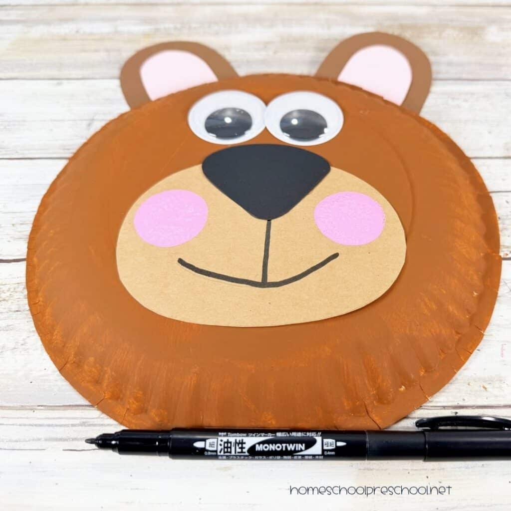 Finished bear paper plate craft with googly eyes, pink cheeks, and a hand-drawn smile on a painted brown paper plate.