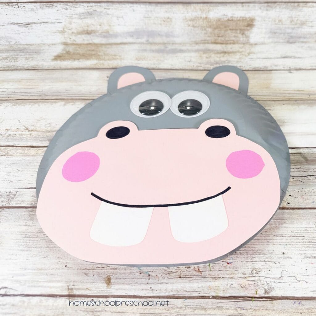 Finished paper plate hippo craft with gray face, pink muzzle and cheeks, wiggle eyes, black nostrils, and two large white teeth.