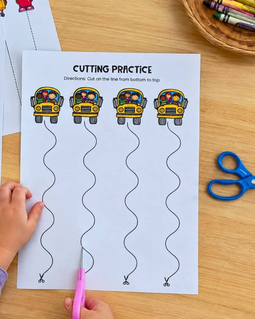 Preschooler cutting along wavy lines on a Back to School cutting practice worksheet featuring school buses.