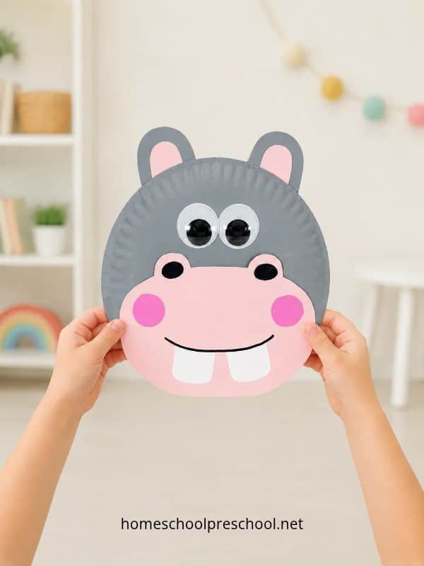 Child holding a finished paper plate hippo craft with a gray face, pink muzzle, wiggle eyes, and white teeth.