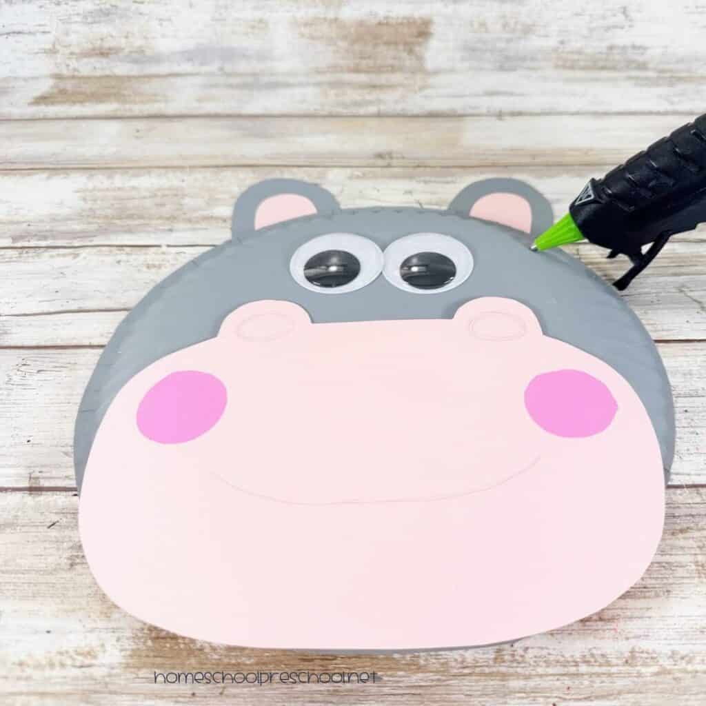 Paper plate hippo being assembled with a pink muzzle, gray and pink ears, and large wiggle eyes.