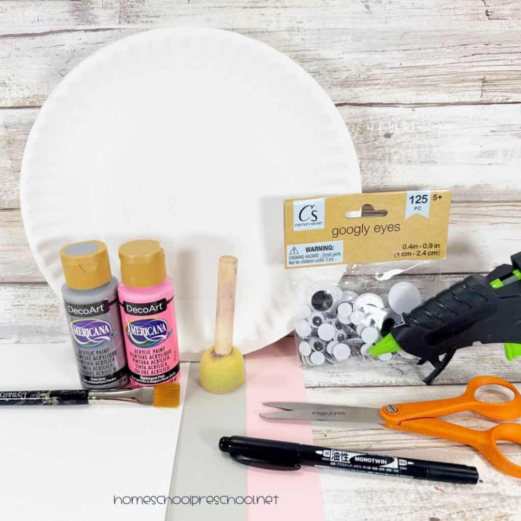 Supplies for a paper plate hippo craft, including a paper plate, gray and pink paint, cardstock, wiggle eyes, scissors, glue, and markers.