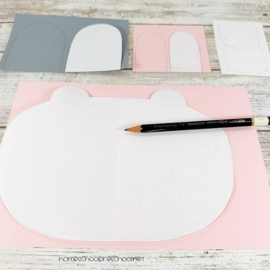 Printable hippo craft patterns being traced onto pink, gray, and white cardstock with a pencil.