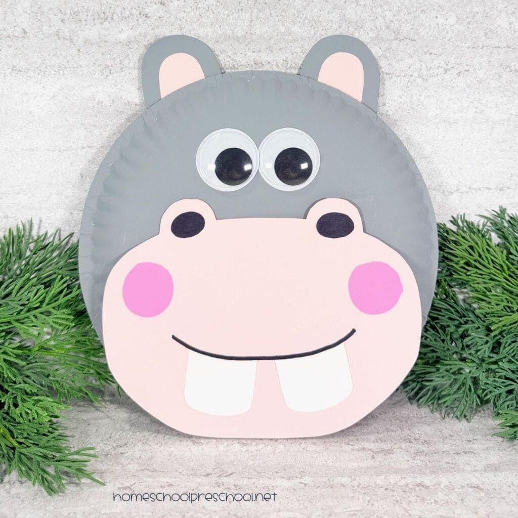 Paper plate hippo craft with gray paint, pink cardstock muzzle and ears, large wiggle eyes, pink cheeks, and white teeth.