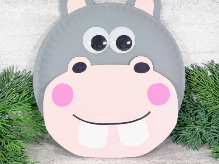 Paper plate hippo craft with gray paint, pink cardstock muzzle and ears, large wiggle eyes, pink cheeks, and white teeth.