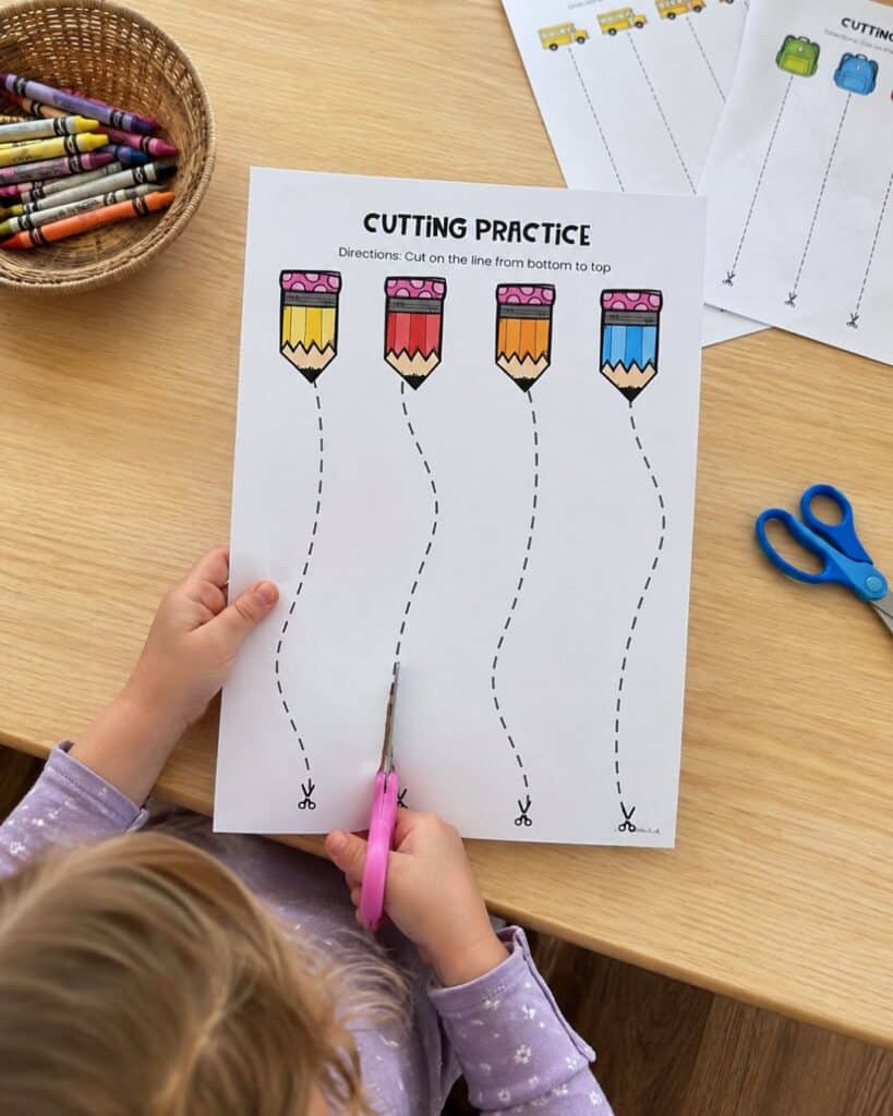 Preschooler using scissors to follow curved dashed lines on a Back to School cutting practice worksheet with colorful pencils.