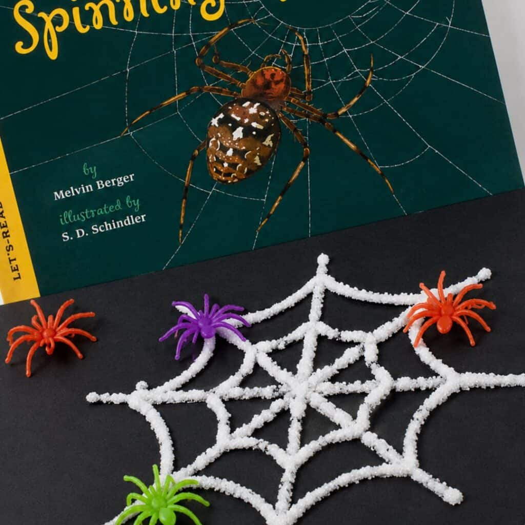 Salt-painted spider web craft with colorful plastic spiders on black paper, paired with the book Spinning Spiders.