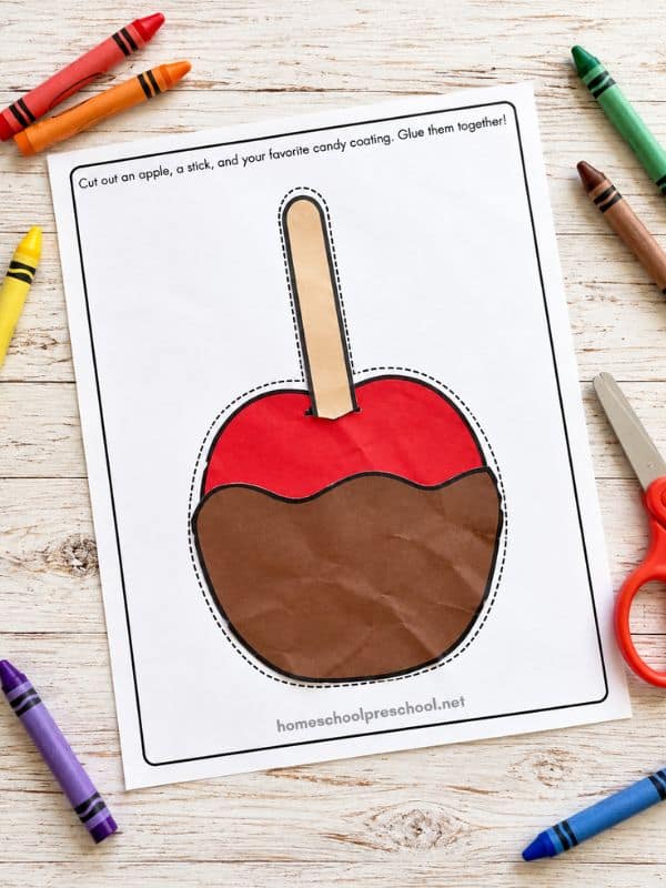Completed cut-and-paste candy apple craft for kids featuring a red paper apple with brown candy coating and a tan stick, surrounded by crayons and scissors.