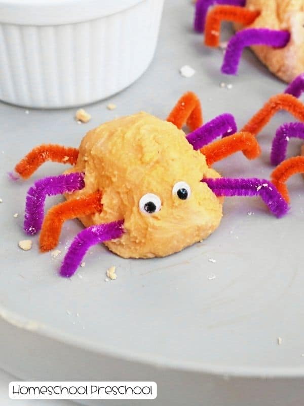 Orange baking soda spider with purple and orange pipe-cleaner legs and two googly eyes on a tray.